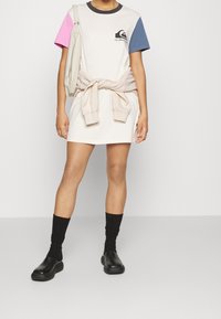 Abito a maniche corte in cotone beige con dettagli rosa e blu, caratterizzato da una scollatura rotonda e un logo. Abbinato a calzini neri e sabot.