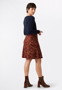 Marineblaues Langarmoberteil, kombiniert mit einem burgunderfarbenen Rock mit gelbem und grünem Muster. Das Model trägt glänzende braune Stiefeletten mit Blockabsatz.