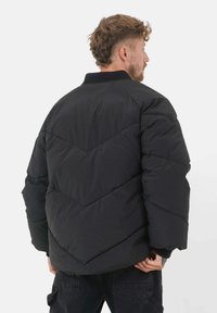 Svart polstret jakke med et skjermønster, med høy krage og lange ermer. Laget av et glatt, lett materiale.