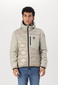 Giacca puffer in beige chiaro con motivo trapuntato, chiusura frontale con zip, cappuccio regolabile e zip nere. Include tasca sul petto con logo.