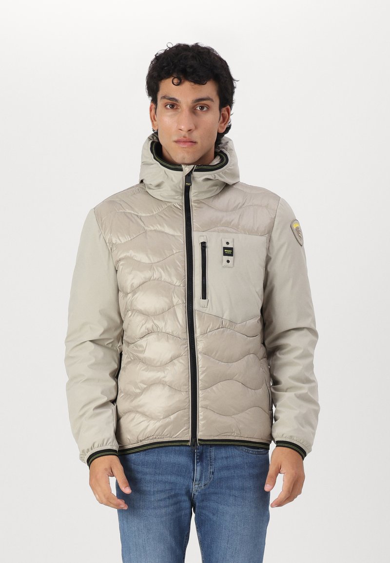 Giacca puffer in beige chiaro con motivo trapuntato, chiusura frontale con zip, cappuccio regolabile e zip nere. Include tasca sul petto con logo.