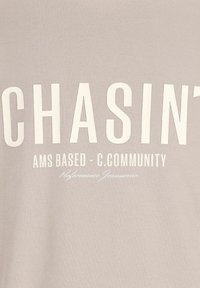 Lys grå bomulds T-shirt med fed hvid tekst: "CHASIN' AMS BASED - C.COMMUNITY" og "Performance Jeanswear" i mindre skrift.