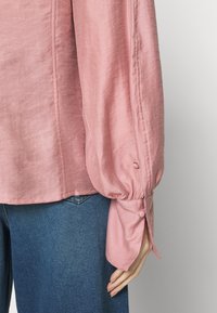 Blouse rose à manches longues avec poignets boutonnés et manches froncées portée avec un jean en denim bleu, montrant une main reposant sur le côté.