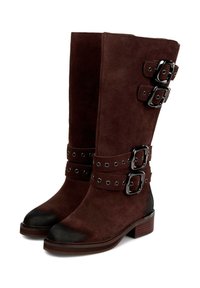 Botas altas de ante marrón con puntera de cuero negro, que presentan hebillas y detalles de ojetes, y un tacón de madera apilada.