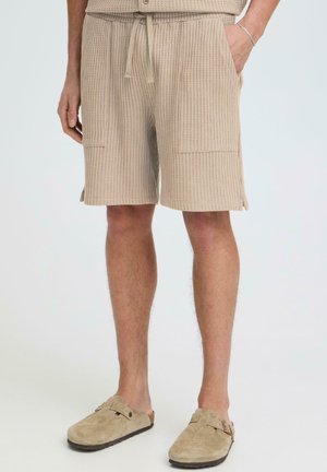 Man wearing beige textured shorts with front pockets and beige slip-on clogs, standing against a plain light background.