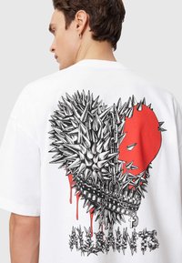 Λευκό T-shirt με μεγάλο γραφικό στο πίσω μέρος: μια καρδιά με αγκάθια σε μαύρο και κόκκινο με σταγόνες και διακοσμητικά αλυσίδες.