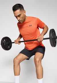 Hombre atlético con una camiseta de manga corta texturizada naranja y pantalones cortos negros, levantando una barra con pesas circulares frente a un fondo gris.