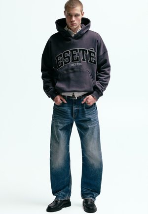 Temno sive barve majica s kapuco z velikimi črkami "ESETÉ ARCHIVES", kombinirana z ohlapnimi, izpranimi modrimi kavbojkami in črnimi čevlji.