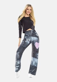 Jauna sieviete ar gariem blondiem matiem, tērpusies melnā garroku krekliņā un izšūtu džinsu biksēs ar rozā sirdi, stāvot pret baltu fona.