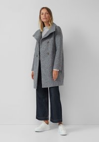 Een vrouw in een grijze dubbelgeknoopte wollen jas over een lichte trui, donkere wijde jeans en witte sneakers, staande tegen een effen muur.