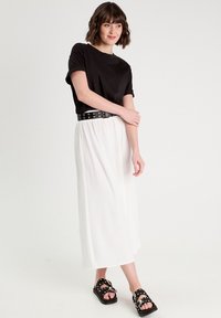 T-shirt noir à manches courtes associé à une jupe maxi blanche et fluide. La tenue est complétée par une ceinture cloutée noire et des sandales à glisser noires avec des accents.