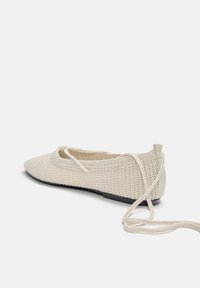 Beige gestrickte Ballerinas mit spitzer Zehe, strukturiertem Obermaterial und schlanker schwarzer Sohle. Mit Schnürbändern an der Ferse für eine verstellbare Passform.