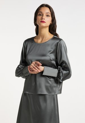Young woman with dark wavy hair wears a shiny gray long-sleeve blouse and matching skirt, standing against a plain white background.