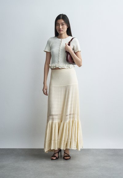 Frau trägt ein cremefarbenes kurzärmeliges, geknöpftes Oberteil und einen langen plissierten Rock, hält eine dunkelburgunderfarbene Schultertasche und steht auf einem grauen Boden.