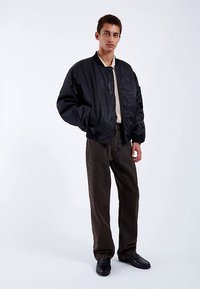 Musta bomber-takki, jossa on sileä pinta, vetoketju edessä ja resorit hihansuissa, yhdistetty tummanruskeisiin chinoihin ja mustiin kenkiin.