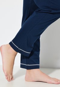 Navy bomullbyxor med lös passform, med ett vitt prickmönster vid fållen, visade bredvid en bar fot på en ljus yta.