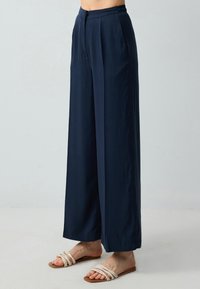 Marineblauwe wijde broek met een gladde textuur, voorzien van een geplooide voorkant en een elastische tailleband. Gepaard met sandalen met bandjes.