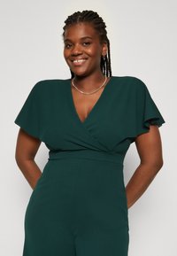 Groene jumpsuit met vouwmouwen, V-hals en strakke taille. Glad, gestructureerd stof met een slank ontwerp. Accent met een zilveren ketting halsketting.