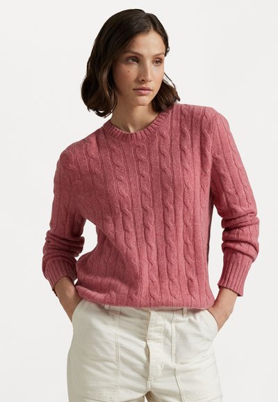 Suéter de punto de cable de color rosa suave con cuello redondo, puños y dobladillo acanalados, hecho de una tela suave, combinado con pantalones de color claro.