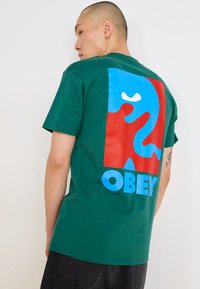 Tricou verde cu mânecă scurtă, având un grafic mare în nuanțe de albastru și roșu cu un chip stilizat și cuvântul "OBEY" scris cu litere îngroșate albastre.