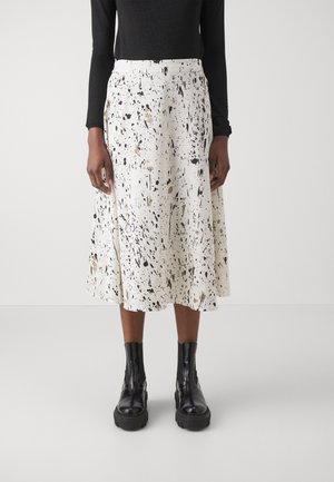 Jupe midi blanche avec motifs éclaboussés noirs et or, en tissu léger ; comporte une coupe évasée et une ceinture ; portée avec des bottes noires.