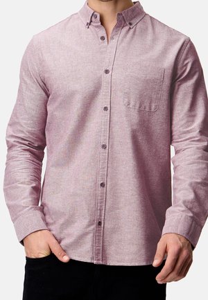 Homme portant une chemise rose à manches longues avec une poche poitrine, une main dans la poche d’un pantalon noir, sur fond clair.