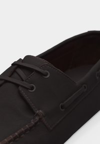 Σκούρο καφέ δερμάτινο μοκασίνι με δέσιμο κορδονιών και ορατή ραφή κατά μήκος της επάνω άκρης, εμφανίζεται σε ανοιχτό φόντο.