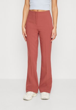 Woman wearing high-waist, wide-leg rust-colored pants paired with white sneakers and a white cropped top, standing against a white background.