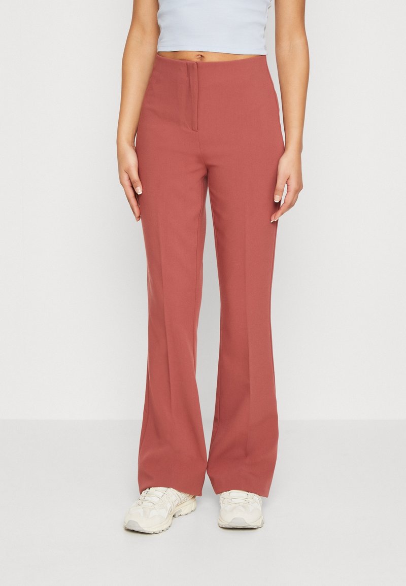 Femme portant un pantalon taille haute à jambes larges de couleur rouille, associé à des baskets blanches et un top court blanc, debout devant un fond blanc.