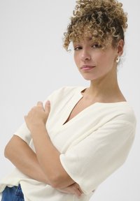 Crème kortemouwen top met een V-hals, gestructureerde stof en een relaxte pasvorm. Model heeft de armen gekruist en staat tegen een effen achtergrond.