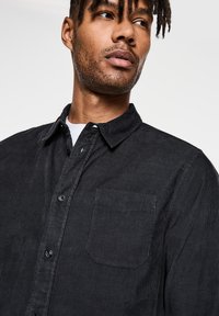 Svart kordfläskjorta med knäppning, bröstficka och vertikala ränder, som har synliga knappdetaljer och en texturerad tyg.