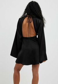 Robe en satin noir avec de longues manches cloche, dos ouvert et taille élastique. Présente une fermeture à nouer au cou. Texture lisse et design classique.