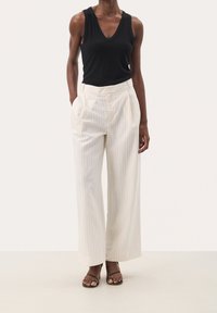 Haut noir sans manches avec un col en V associé à un pantalon large beige à fines rayures verticales. Le tissu semble lisse avec une coupe décontractée.