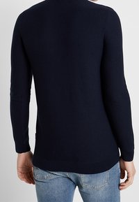 Mörkblå stickad tröja med långa ärmar och ribbad nederkant, med en slät yta och minimalistisk design, som bärs över ljusa jeans.