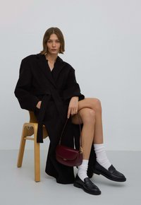 Zwarte wollen jas met een riem, gecombineerd met zwarte loafers en witte sokken. Bourgondische handtas voegt kleur toe. Model zittend op een houten stoel.