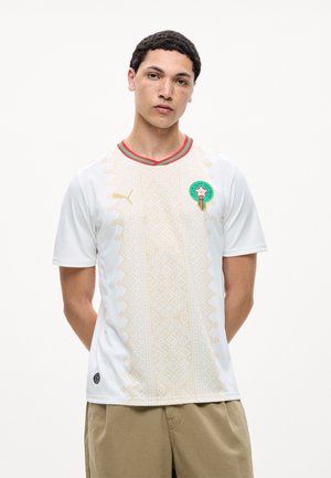 Jonge man draagt een wit Marokko voetbalshirt met goudkleurig patroon, rode en groene halslijn, staand tegen een effen witte achtergrond.