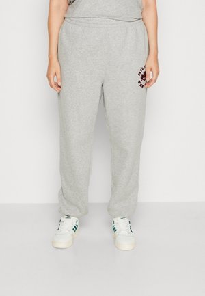 Osoba nosząca jasnoszare spodnie dresowe z małym logo na lewym udzie oraz białe sneakersy Adidas z zielonymi paskami.
