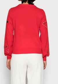 Rode trui met geribde zoom en ballonmouwen. Voorzien van geborduurde accenten bij de manchetten en een ronde halslijn. Gemaakt van gebreide stof.