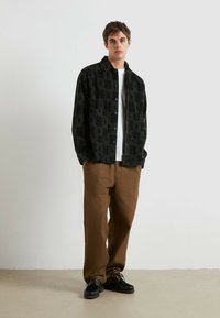 Giacca nera con motivo circolare verde, t-shirt bianca sotto, pantaloni marroni e scarpe nere con lacci e suole testurizzate.