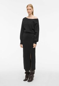 Črna pletena dolga obleka z off-the-shoulder izrezom in dolgimi rokavi. Ima stranski razporek. V kombinaciji z visokimi temno rjavimi čevlji.