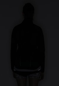 Zwarte sportjack met hoge kraag, reflecterende accenten dichtbij de zoom en een aansluitend ontwerp. Draagt over een geblokte short.