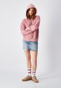 Sudadera con capucha rosa de corte relajado, combinada con una falda de mezclilla desgastada. Zapatillas de deporte color beige y calcetines blancos con rayas rojas completan el look.