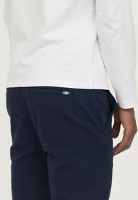 Navyblauwe broek van een gestructureerde stof, met een achterzak met een label en zijzakken; gecombineerd met een witte langslaven blouse.