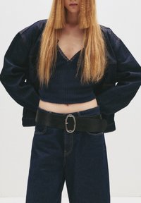 Temno jeans jakno in hlače z visokim pasom v kombinaciji z mornarsko modrim kratkim topom z čipkastim robom, dopolnjeno s širokim črnim pasom s srebrno zaponko.