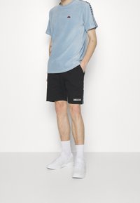 T-shirt en velours côtelé bleu clair avec col rond ; short noir avec logo latéral et bande multicolore ; chaussures de sport blanches et chaussettes.