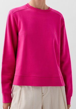 Persoon draagt een felroze sweater met lange mouwen en ronde hals en beige broek met hoge taille tegen een effen achtergrond.