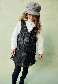 Ein junges Mädchen trägt eine graue Mütze, ein weißes Langarmshirt, ein schwarzes Kleid mit floralen Strukturen, schwarze Leggings und schwarze Stiefel vor einem gedämpften Hintergrund.