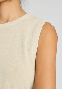 Beige bezramínkový top s žebrovaným výstřihem a texturovaným pleteným materiálem, který ukazuje hladký, přiléhavý siluetu.