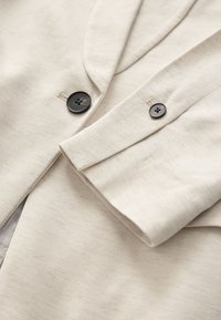 Beige stof met een gestructureerde afwerking, voorzien van twee donkere knopen en een omgevouwen kraag. Close-up benadrukt materiaal- en stiksel details.