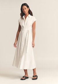 Witte overhemdjurk met korte mouwen, een ceintuur in de taille en een vloeiende rok tot aan de enkels. Gecombineerd met zwarte slippers.